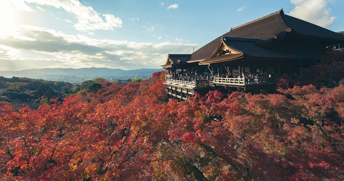 A Japanese temple framed by bright red autumn maple leaves under a clear blue sky